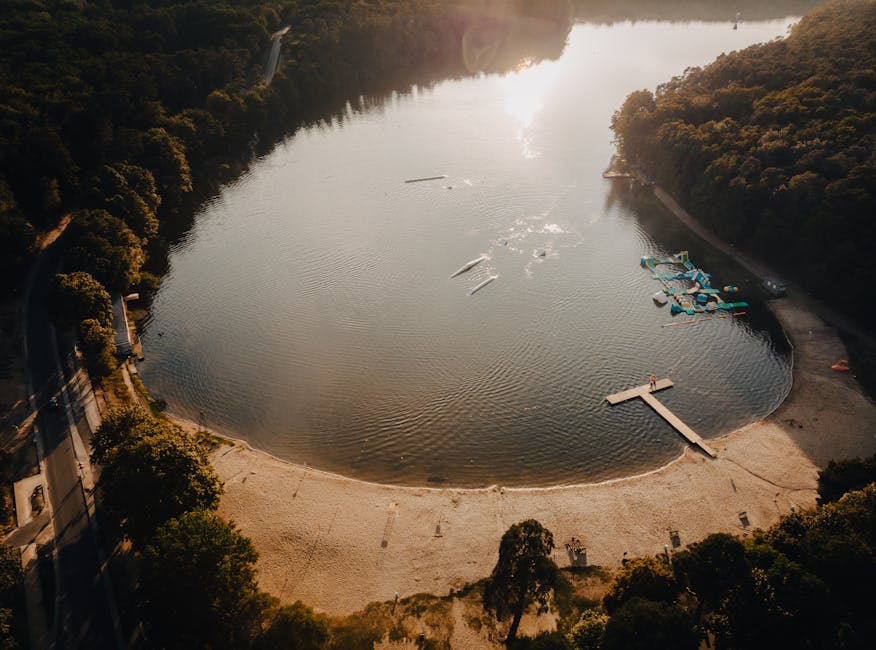 An aerial view of a small, circular embayment at Ruislip Lido, showing a sandy beach area along the shoreline with a wooden jetty extending into the calm water. The water surface reflects the sunlight, creating a bright, shimmering effect, while several small boats and watercraft are visible, some moving across the water. Surrounding the bay are lush, green trees that line the edge of the shoreline and extend into the hilly landscape beyond. To the right, a section of inflated water slides and play structures are arranged on a dedicated area adjacent to the beach, with a few people nearby. A narrow road runs along the top of the landscape, bordered by trees, and there are visible shadows cast by the trees and structures, indicating a setting sun. The scene depicts a peaceful, natural environment with recreational facilities, aligning with outdoor waste management and disposal considerations carried out by services like Rubbish Collection Ruislip, particularly relevant in maintaining cleanliness around public leisure areas at sites such as Ruislip Lido.