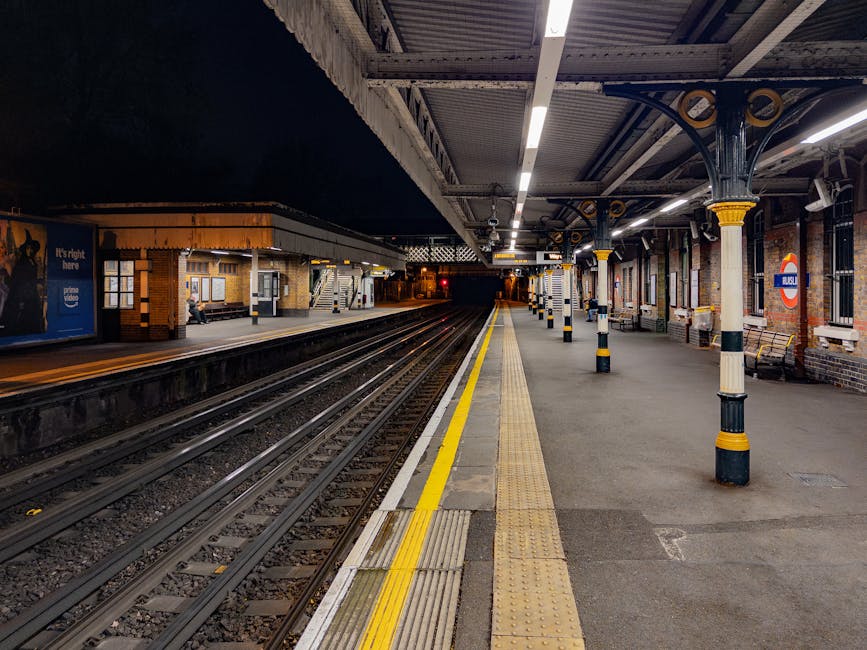 A nighttime view of a railway station platform showing a row of evenly spaced black and cream-colored columns supporting a metal canopy with decorative yellow accents. The platform surface is paved with concrete and features tactile paving strips for accessibility, alongside a yellow safety line near the train tracks. The station includes benches, small brickwork buildings, and advertising boards, with some seating and benches visible along the edge. On the far side of the tracks, there are additional station structures and a person sitting on a bench under warm lighting, indicating a quiet period with minimal activity. The station is well-lit with overhead fluorescent lights, and a London Underground roundel sign is visible on one of the columns, suggesting this is an underground or suburban rail station. The environment appears clean and organized, with a focus on safety and accessibility for passengers. This image from Rubbish Collection Ruislip shows an urban public transport setting that may be associated with ongoing infrastructure or maintenance work, indirectly related to waste and rubbish management needs for station upkeep or construction projects.