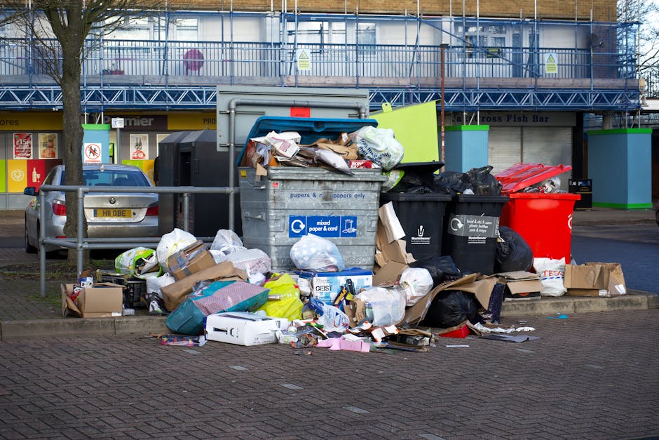 An outdoor scene showing multiple overflowing rubbish bins and collection points on a paved area, with black, red, and black recycling bins filled with discarded cardboard, paper, plastic, and packaging materials. Some waste items, including flattened cardboard boxes, plastic bags, and crumpled paper, are spilling onto the surrounding ground. Behind the bins, there is a metal railing, a parked grey hatchback car, and a building with a storefront featuring yellow signage. In the background, a two-story structure is visible, covered in blue scaffolding and construction netting, indicating ongoing building work. The scene appears to be in a commercial or urban environment during daylight, with natural light illuminating the cluttered waste collection point, exemplifying typical private rubbish disposal practices separate from municipal refuse services, aligned with independent rubbish removal offerings.
